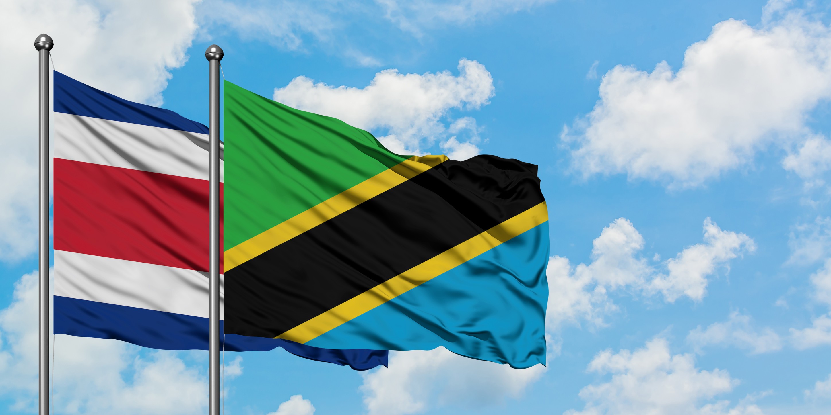 Costa Rica And Tanzania Flag Waving In The Wind Against White Cloudy Blue Sky
