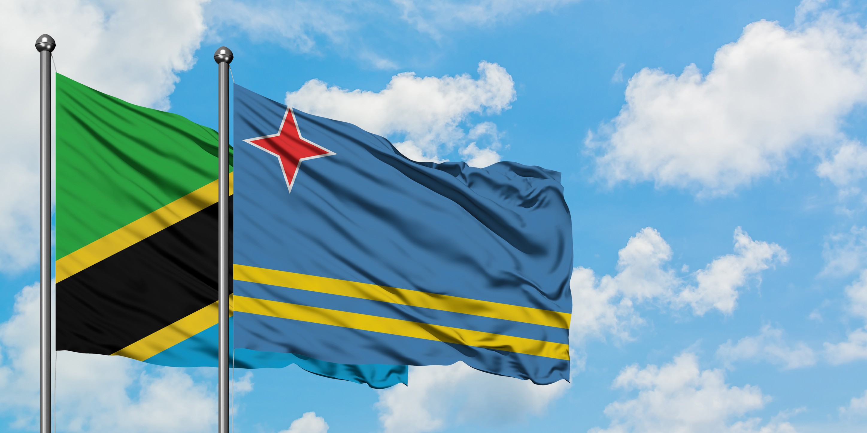 The Flags Of Tanzania And Aruba Wave In The Wind Against A Cloudy Blue Sky