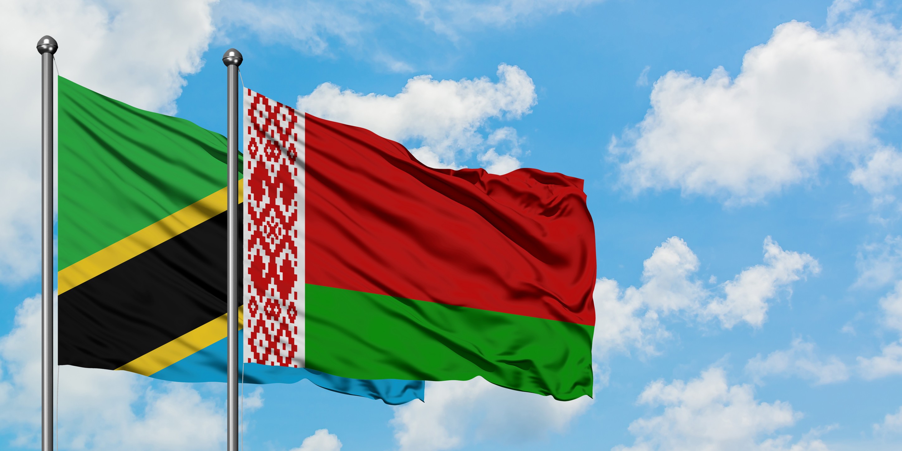 The Flags Of Tanzania And Belarus Wave In The Wind Against A Cloudy Blue Sky